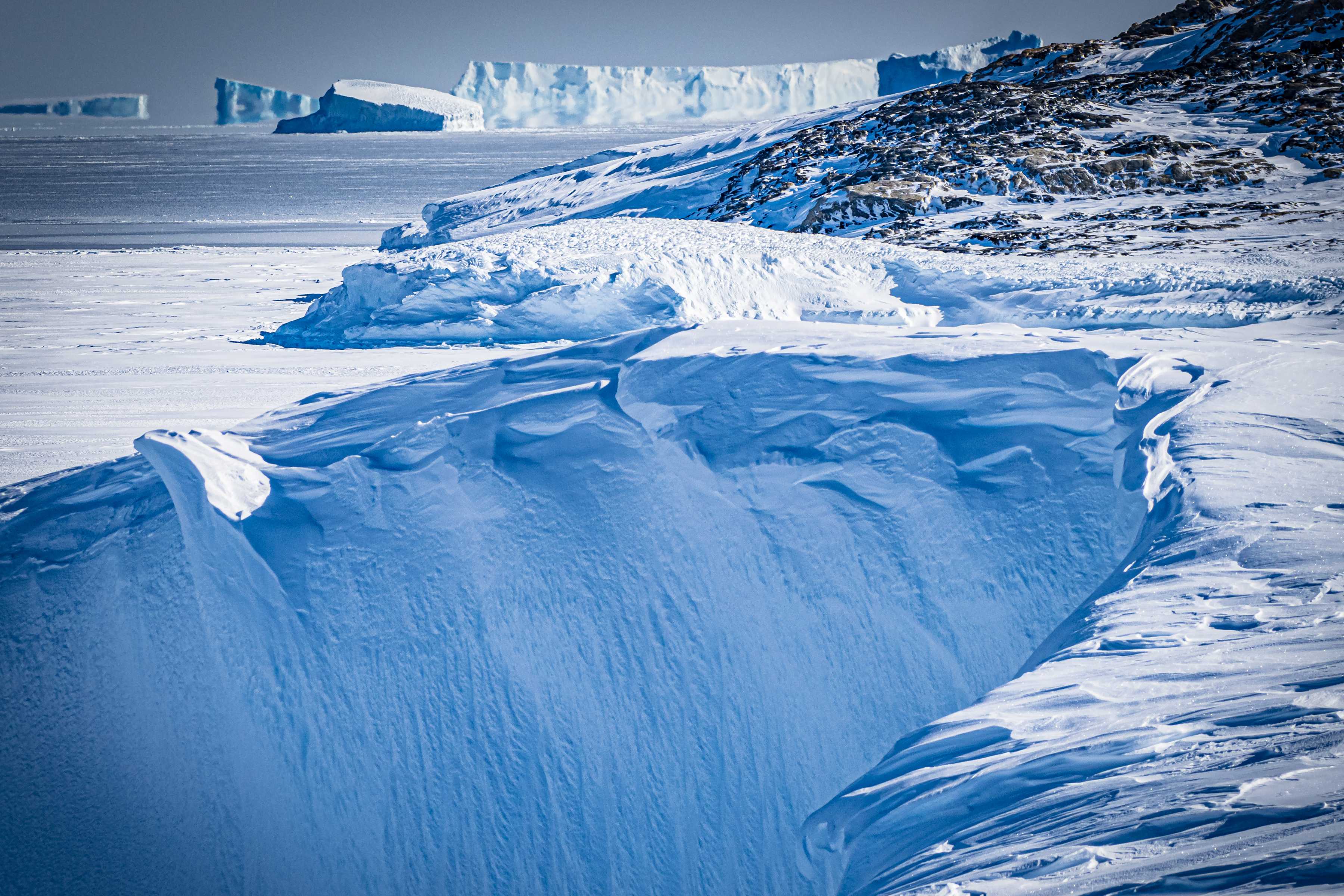 A landscape photo of foreground ice cliff billowed frozen with midground fast-ice edge into open ocean and a backdrop of icebergs. Photo: Sophie Counsell.